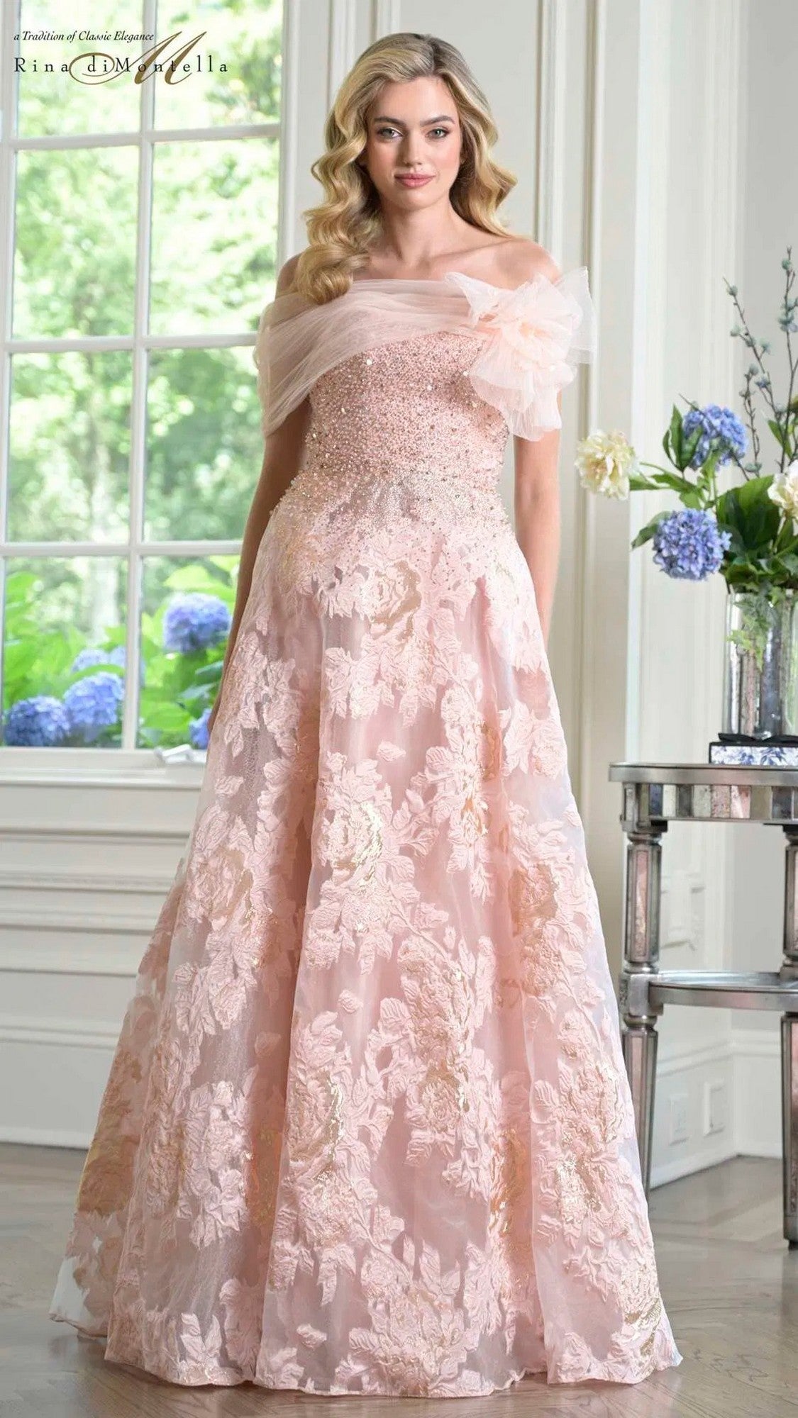 A woman stands indoors by a window and flower-adorned table, wearing the Rina Di Montella RD3406 long formal dress in blush, featuring a beaded bodice and floral embroidery.